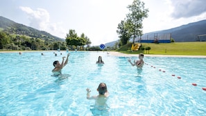 A swimming pool with people in the water, a slide, and a mountainous landscape in the background.