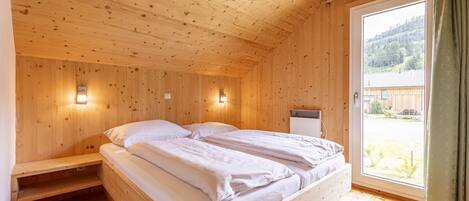 Ein Zimmer in einer Holzhütte mit einem Bett, Nachttischlampen und einem Fenster mit Blick auf einen bewaldeten Bereich.