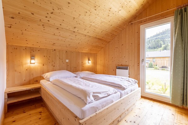 Ein Zimmer in einer Holzhütte mit einem Bett, Nachttischlampen und einem Fenster mit Blick auf einen bewaldeten Bereich.