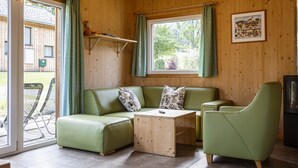 Une pièce chaleureuse avec un canapé vert, une table basse en bois et un fauteuil vert. Il y a une fenêtre avec des rideaux, un tableau encadré au mur et une petite étagère avec un ours en peluche.