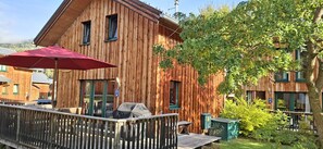 A wooden cabin with a deck, a red umbrella, and a view of the surrounding greenery.