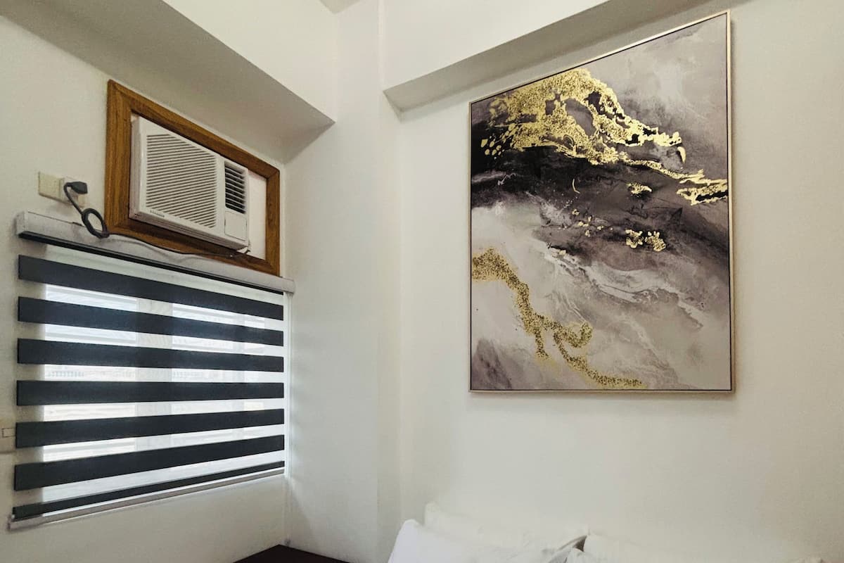 A bedroom with a bed, a window with blinds, an air conditioning unit, and a wall-mounted painting.