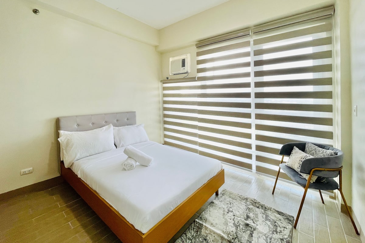 A neatly made bed with white linens and a rolled towel, a grey upholstered chair, and a window with horizontal blinds.