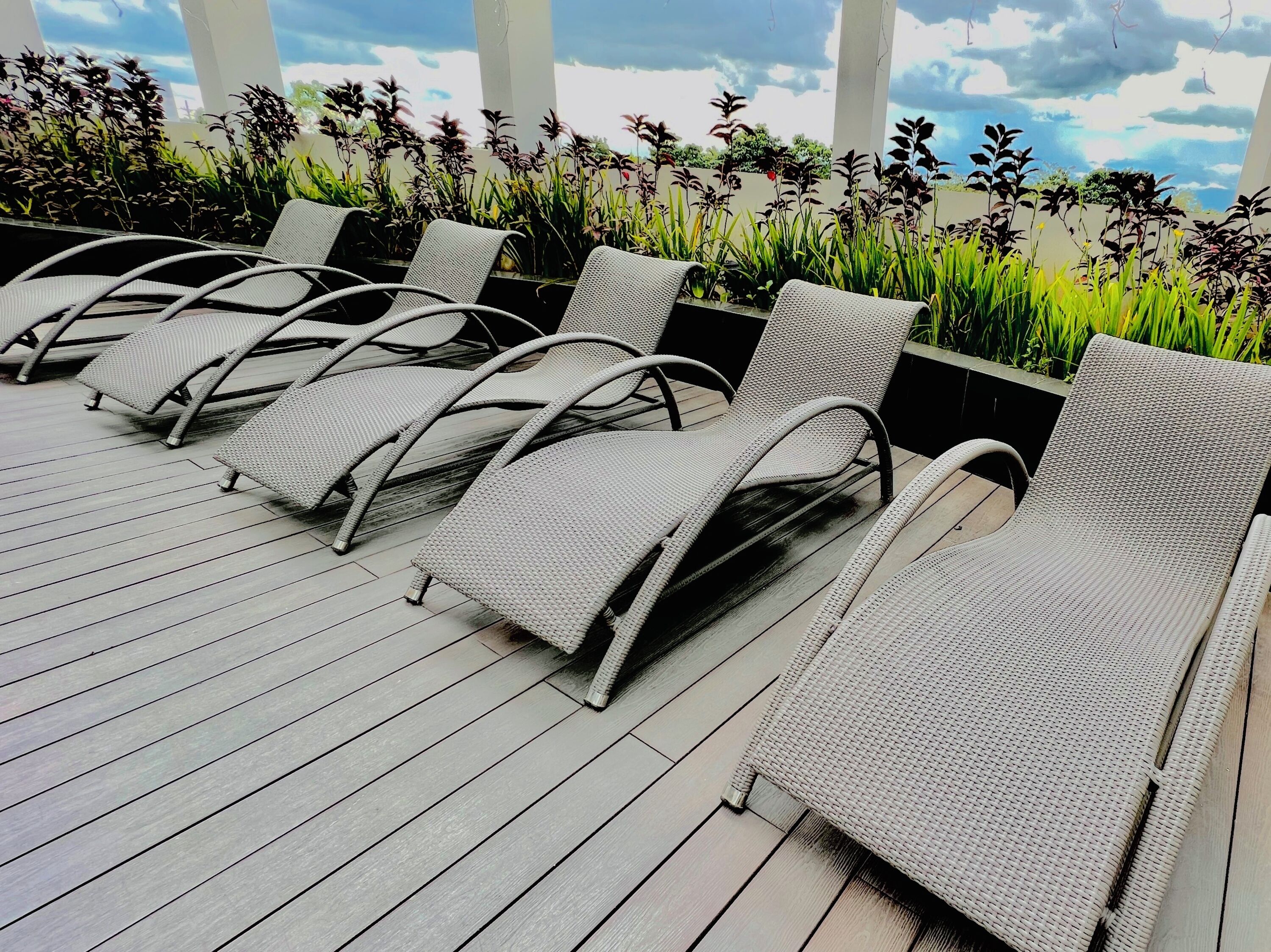A row of wicker lounge chairs on a wooden deck with a view of plants and a cloudy sky.