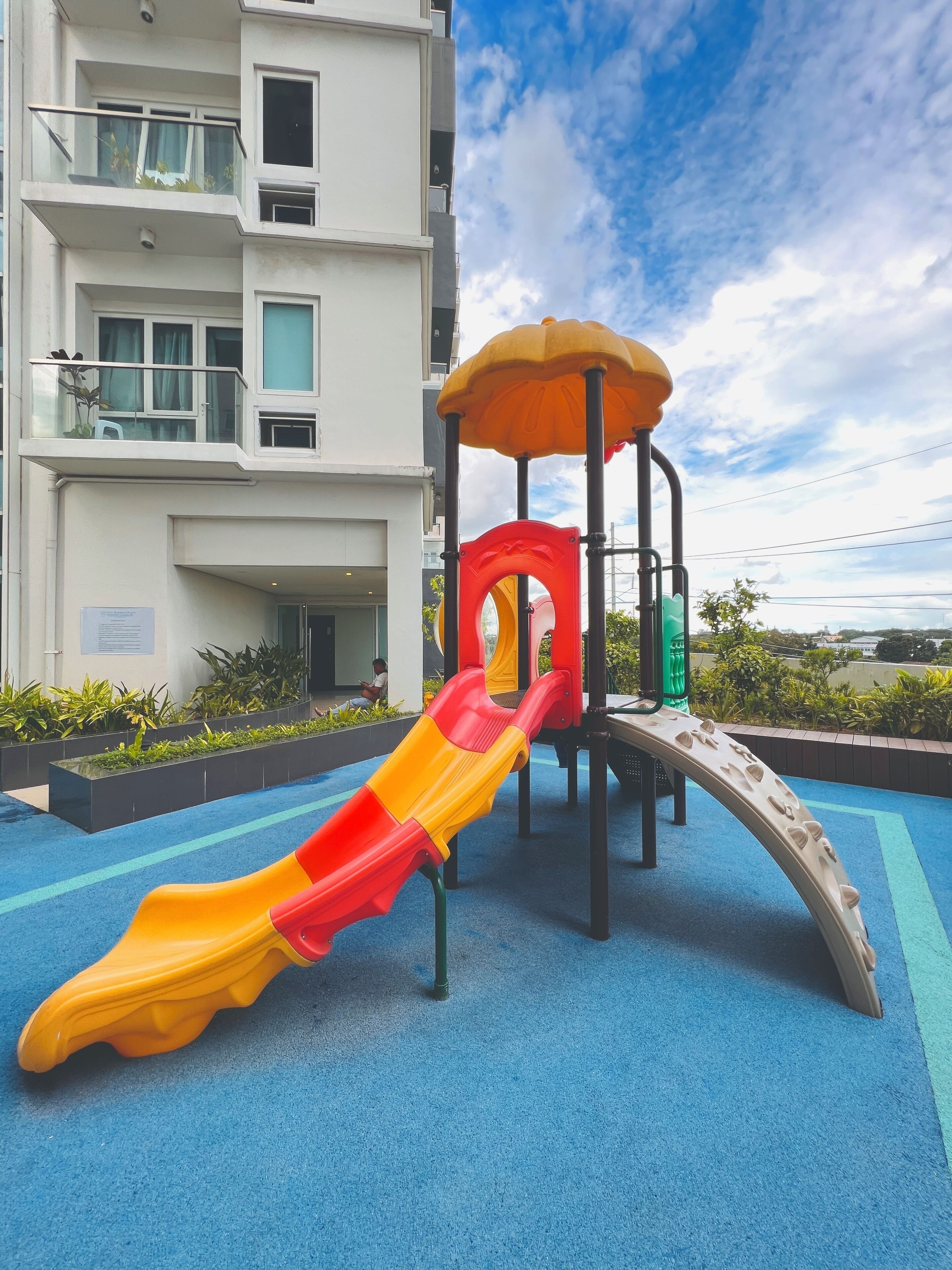 Un parque infantil con un tobogán y estructuras para escalar, ubicado frente a un edificio de varios pisos con balcones.