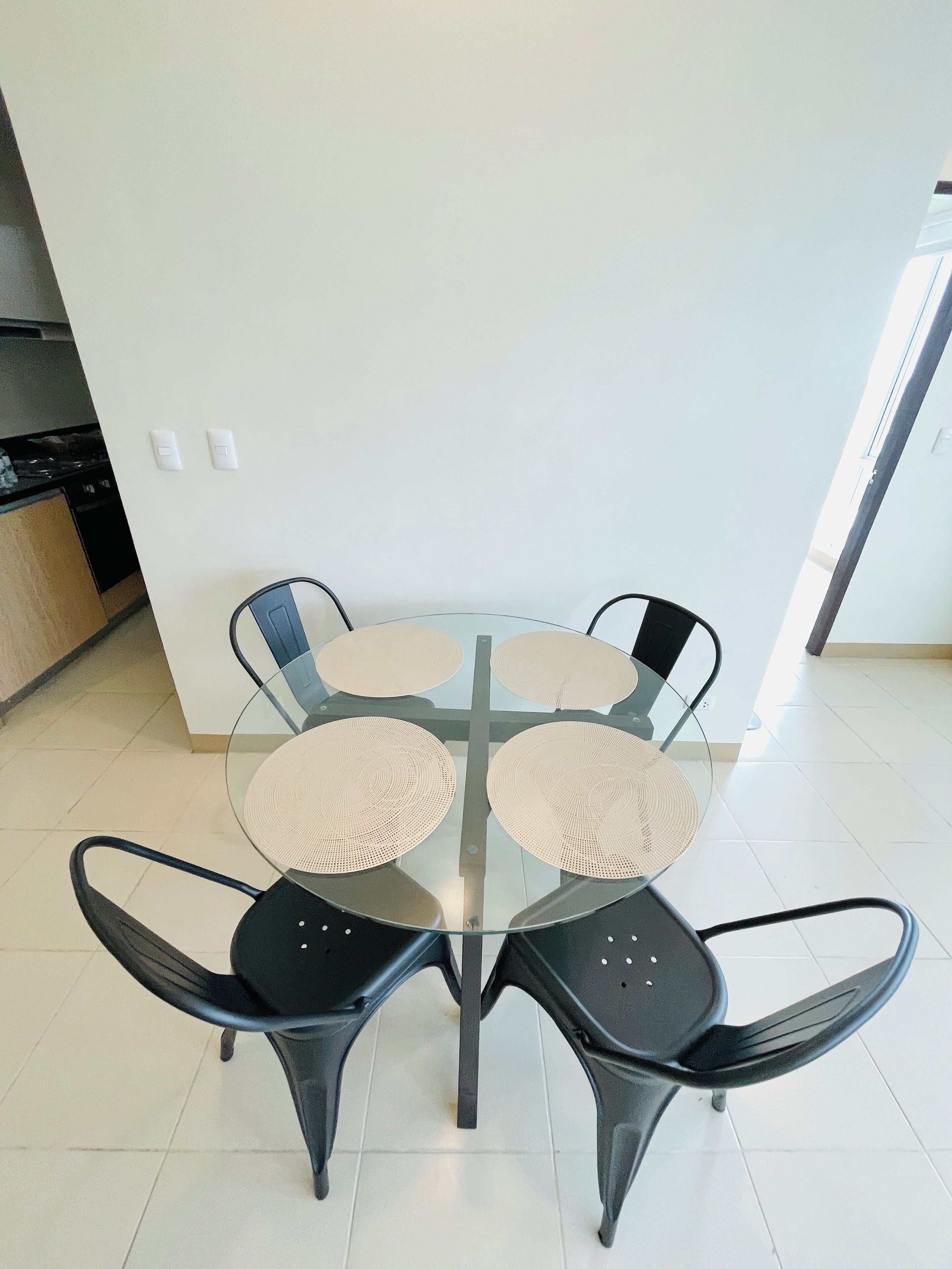A modern dining area with a glass table and four black chairs, featuring woven placemats.