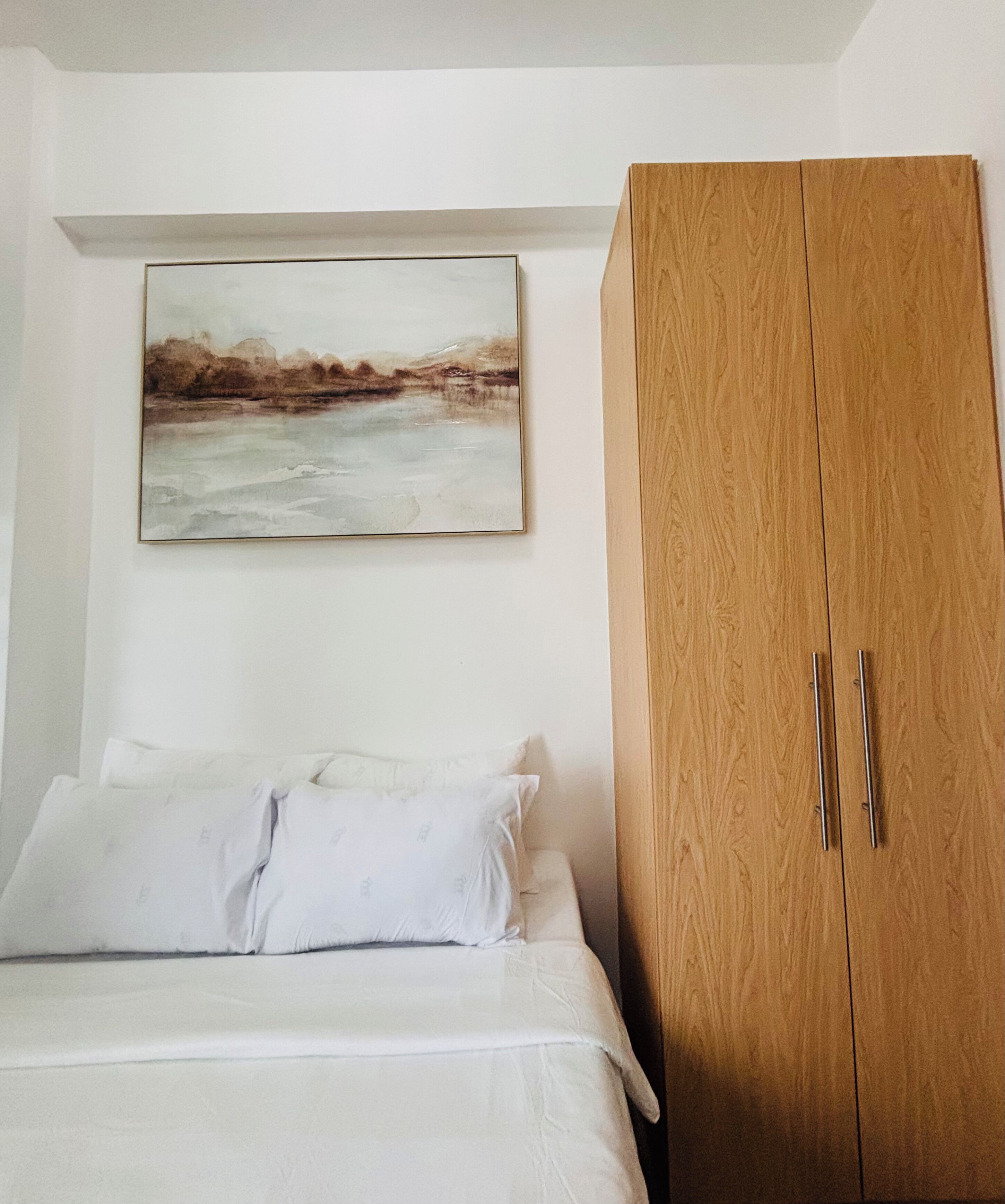Une chambre à coucher avec un lit, du linge de lit blanc, une armoire en bois et un tableau représentant un paysage accroché au mur.