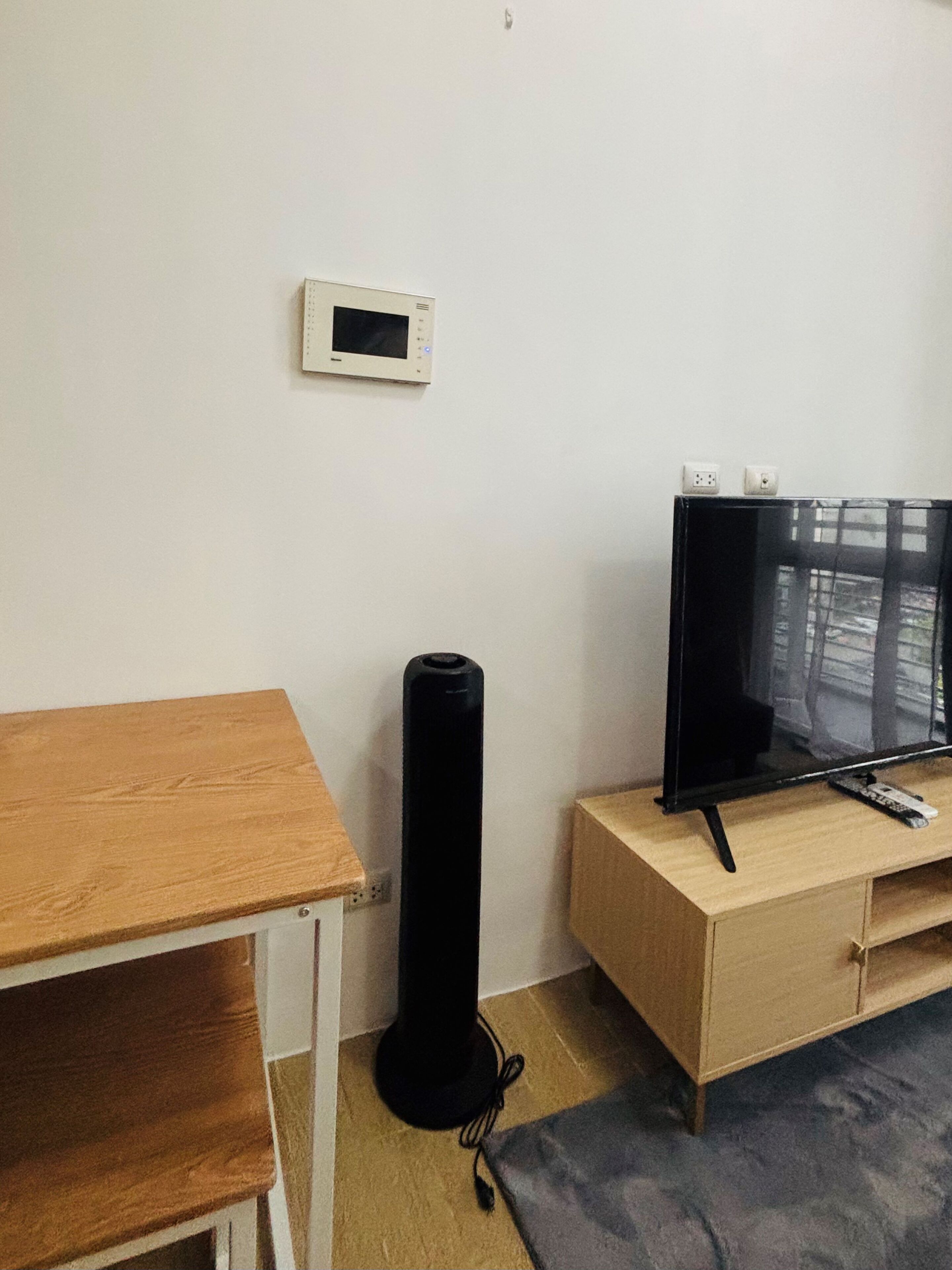 A room with a wooden table, a black speaker, a TV on a wooden stand, and a thermostat on the wall.