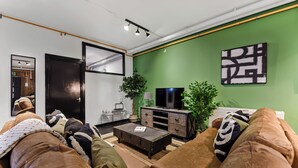 A modern living room with a green accent wall, a large flat-screen TV, a dark wooden coffee table, and a brown leather sofa with patterned cushions. - Trendy, Arty, 3BR in Heart of Hipster Hackney! (London)