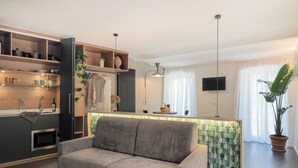 A modern living room with a grey sofa, a kitchen area with wooden shelves, a dining table, and a large potted plant.