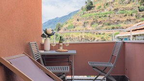 A balcony with two lounge chairs, a table with a vase and wine glasses, and a view of a hillside.