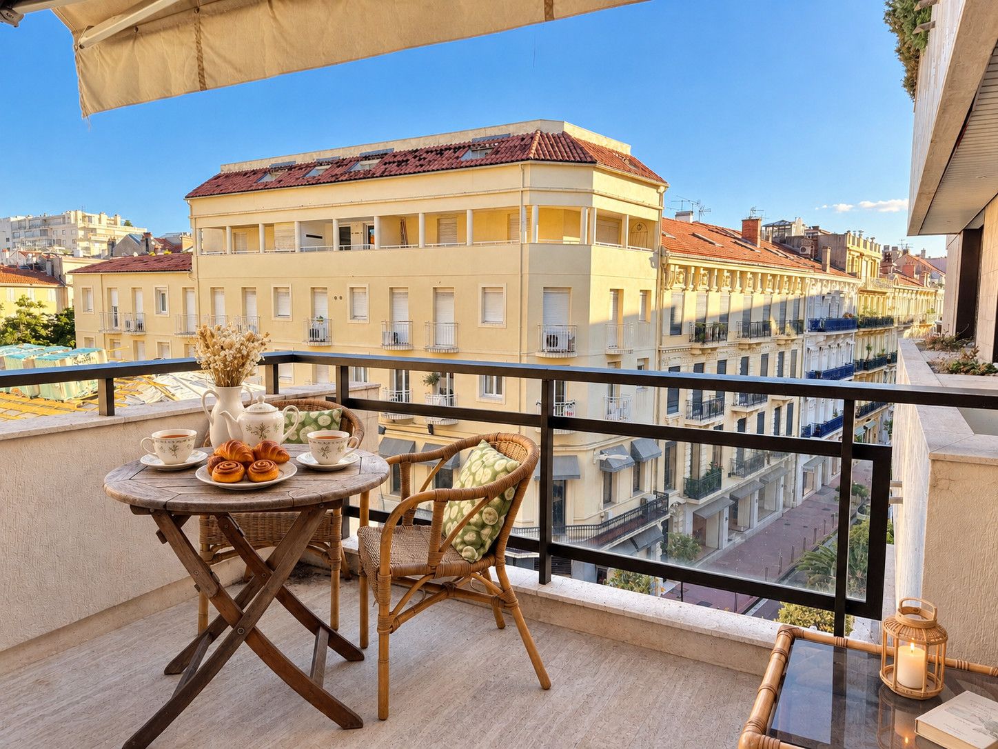 A balcony with a table set for two, a wicker chair, and a view of a building with a red-tiled roof.