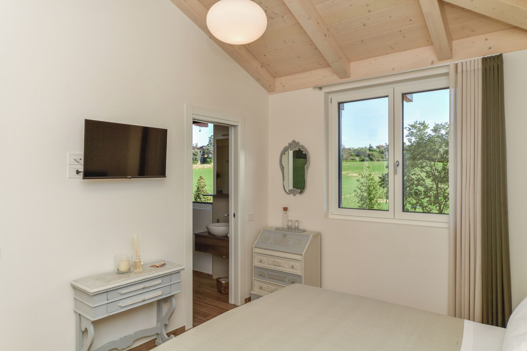 Una camera da letto con un letto, una cassettiera, uno specchio, una televisione e una finestra con vista sul verde.