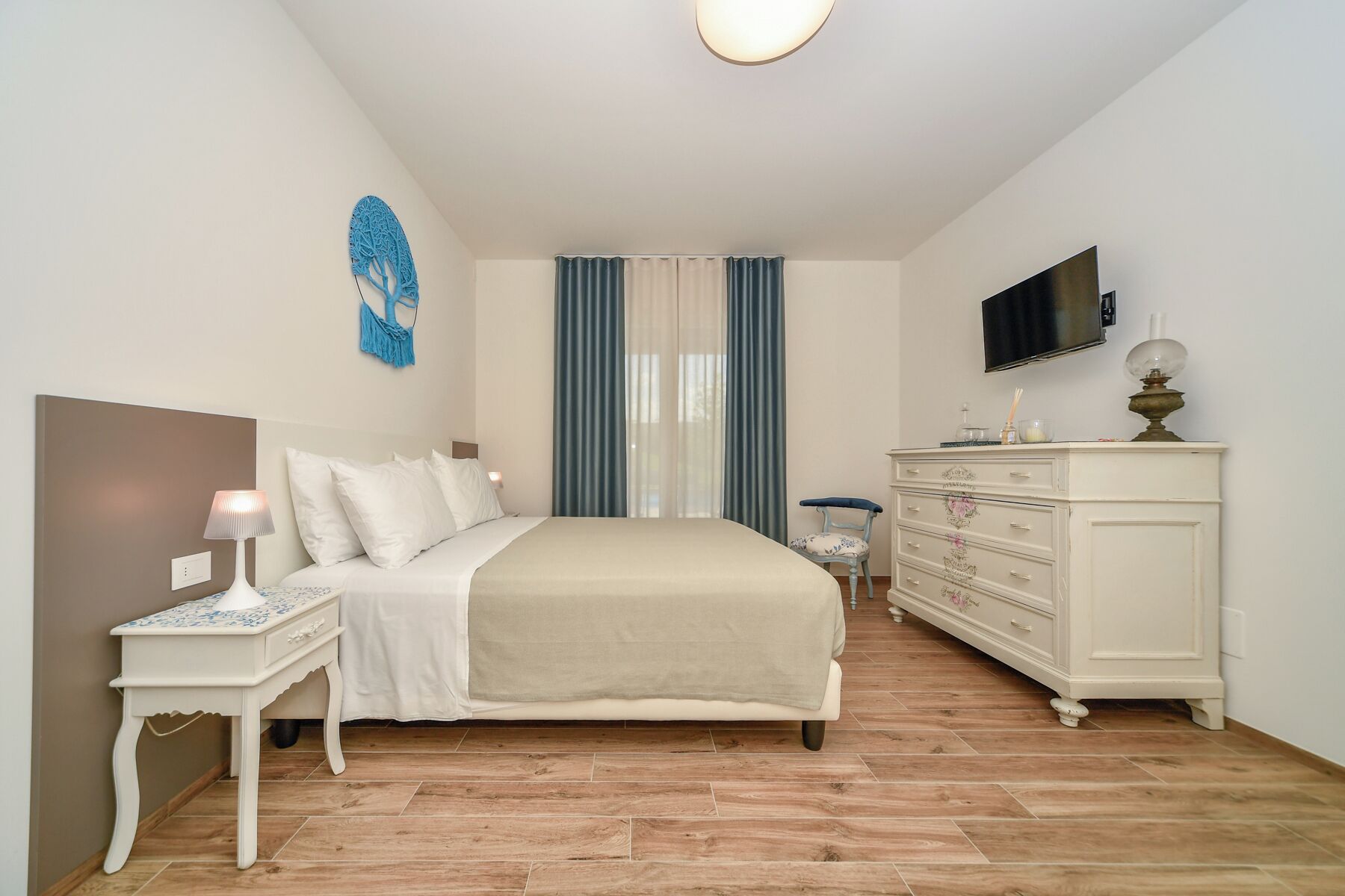 A bedroom with a bed, bedside table, dresser, TV, and curtains.