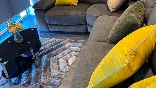 A modern living room with a grey sectional sofa, yellow cushions, a black coffee table with a vase of flowers, and a patterned rug.