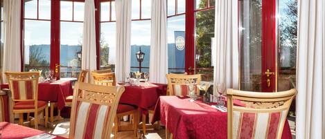 A dining area with tables set for four, wooden chairs, and large windows with red frames.
