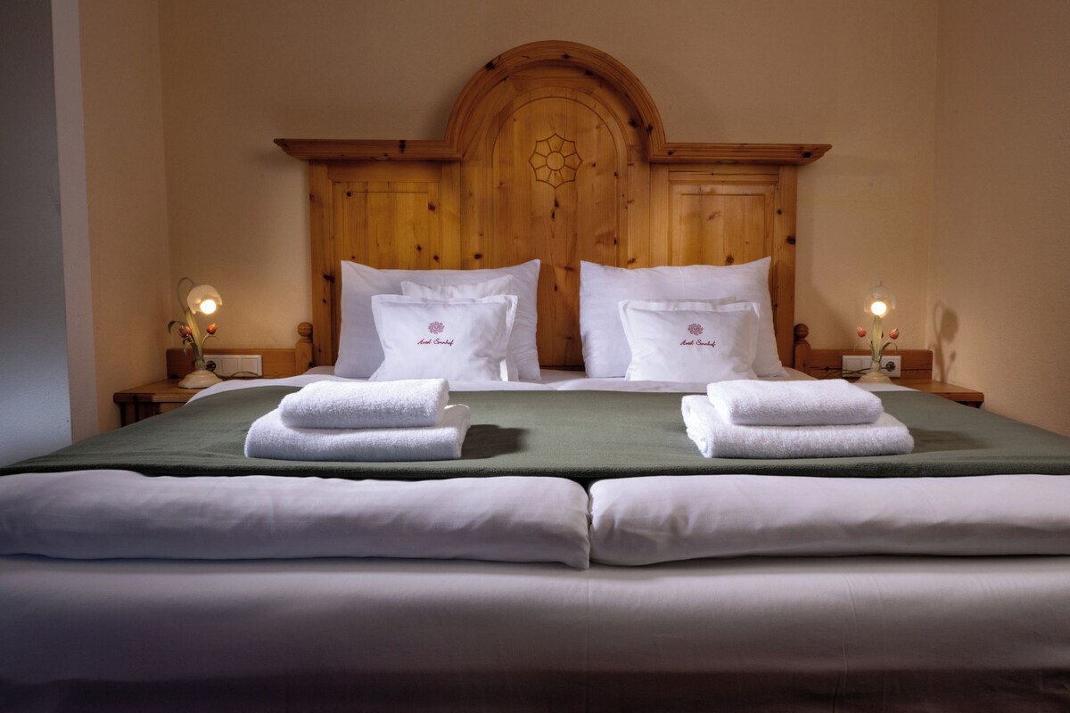 A double bed with white linens, two pillows with a floral design, and a green bedspread. There are two bedside tables with lamps and a decorative vase.