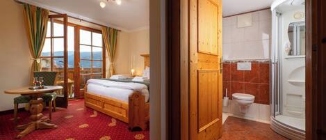 A hotel room with a bed, a desk with a chair, a wooden wardrobe, a bathroom with a shower, and a view of the mountains through the window.