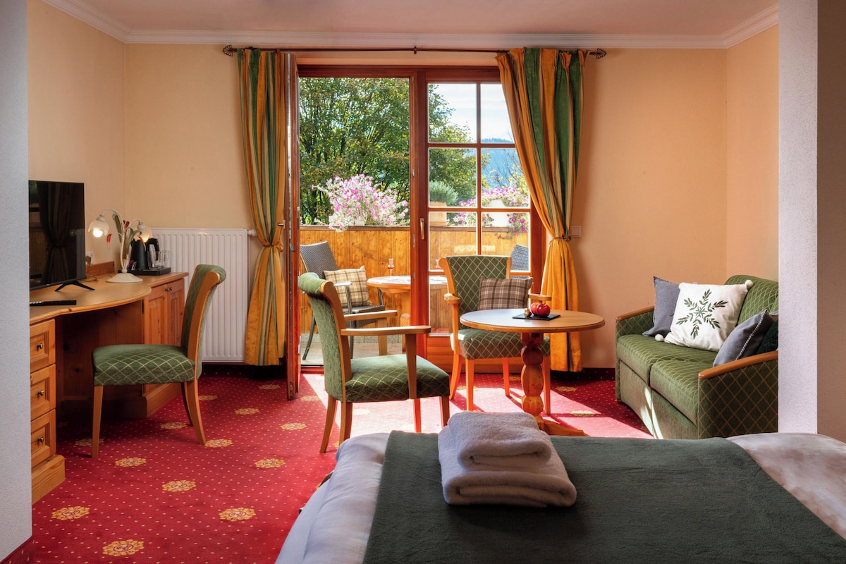 A hotel room with a bed, a desk with a computer, a chair, a small table, and a sofa. There is a window with curtains, and a view of the outdoors.