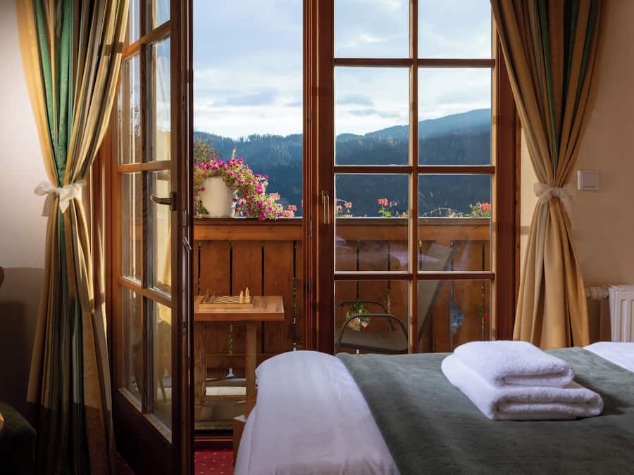 Quarto com cama, uma mesa de madeira e vista para montanhas por uma grande janela.