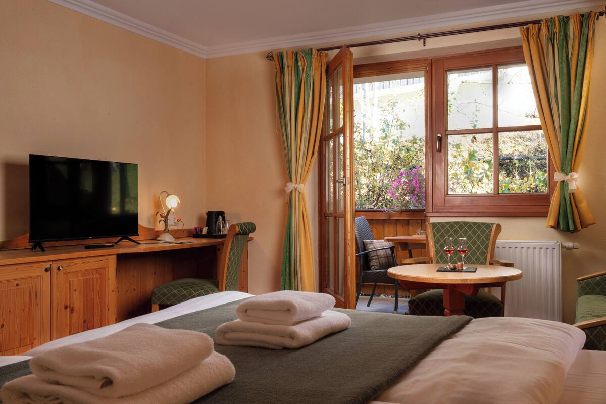 Une chambre d’hôtel avec un lit, une télévision, un bureau avec une lampe, une chaise, une petite table avec des verres à vin et une fenêtre avec des rideaux.