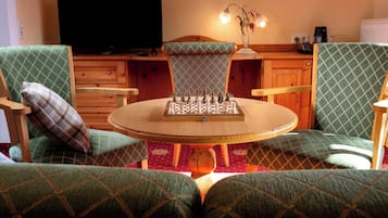 Ein gemütliches Wohnzimmer mit einem runden Holztisch, grün gemusterten Sesseln, einem Schachbrett, einem Fernseher und einer Lampe.