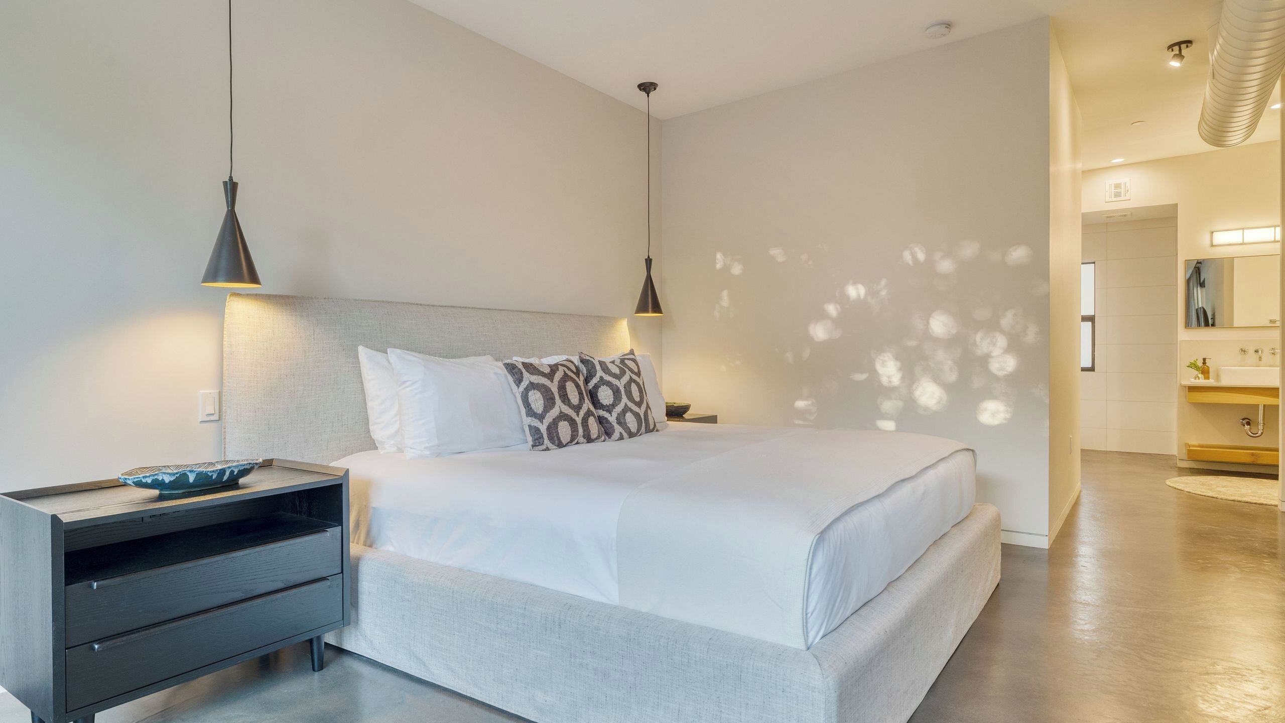 A modern hotel room with a bed, bedside tables, a nightstand, a lamp, and a bathroom visible in the background.
