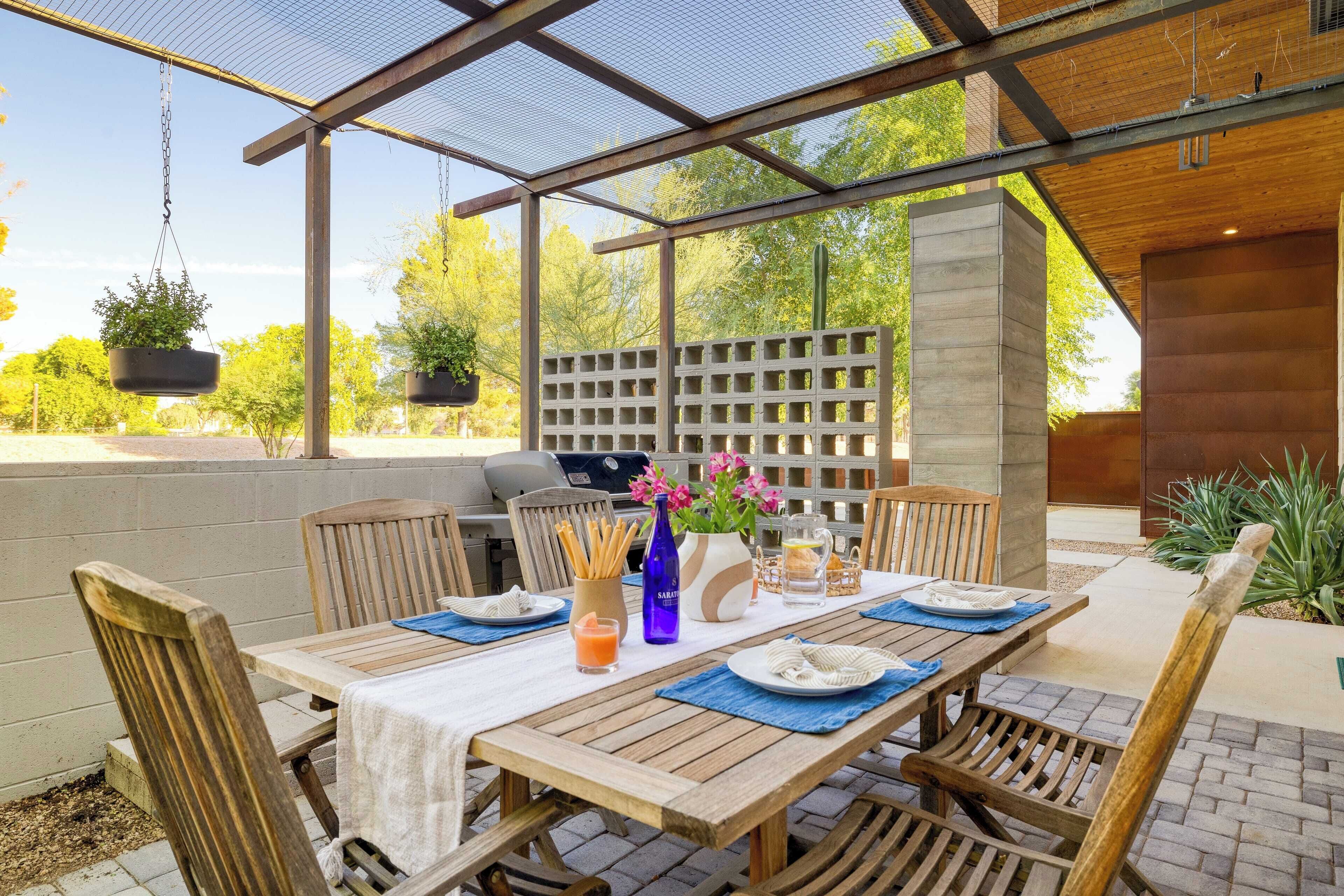 An outdoor patio with a wooden table set for dining, surrounded by chairs, a grill, hanging planters, and a view of trees and a brick wall.