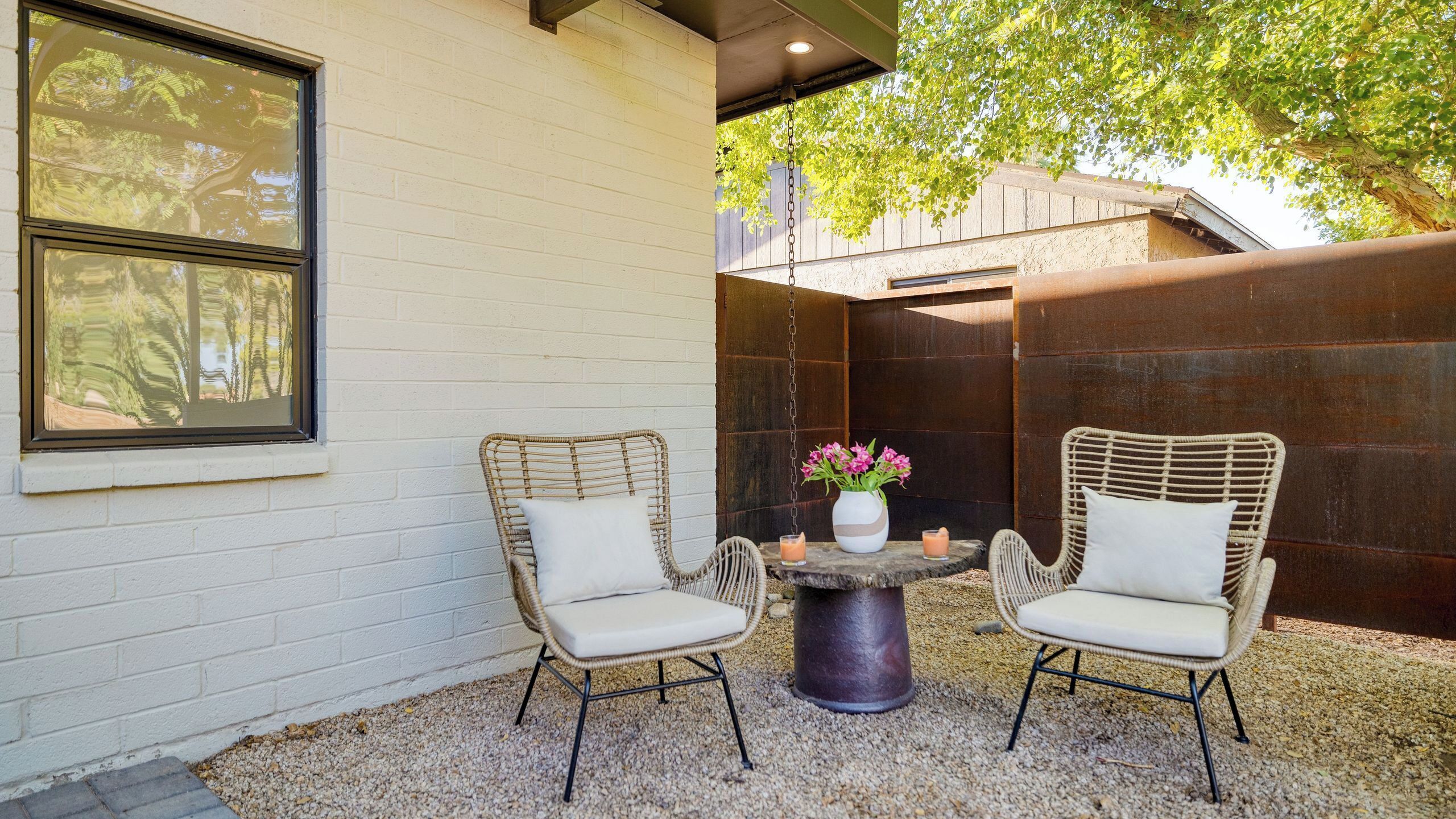 An outdoor seating area with wicker chairs and a table, a white brick wall, and a large window.