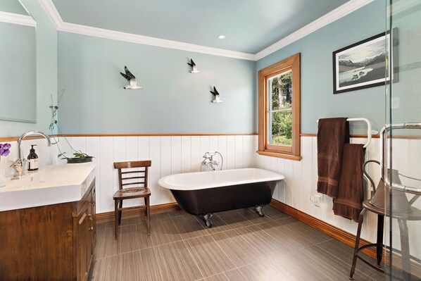 A bathroom with a freestanding bathtub, wooden chair, white vanity, and a window with a view of greenery. - Modernized country house set in beautiful gardens with swimming pool.
(Rangitumau)