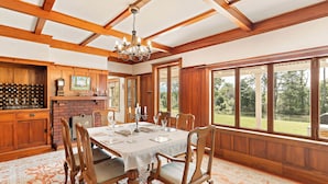 A dining room with a large wooden table, chairs, and a chandelier. There is a brick fireplace and large windows with a view of the outdoors. - Modernized country house set in beautiful gardens with swimming pool.  
 (Rangitumau)