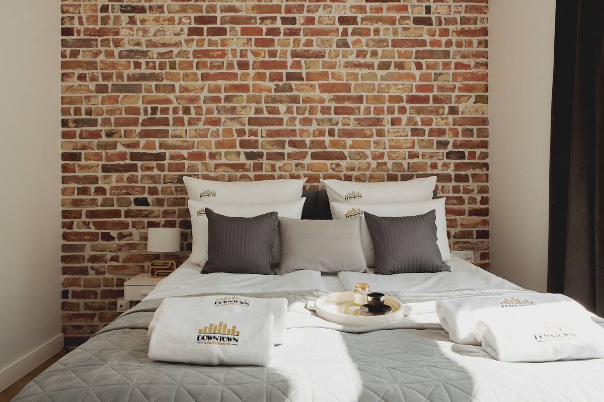 A hotel room with a brick-patterned wall, a bed with white bedding, grey and white pillows, and a tray with a bottle and a bowl.