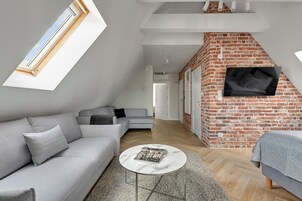A modern loft bedroom with a brick wall, a flat-screen TV mounted on the wall, a grey sofa, and a round coffee table.