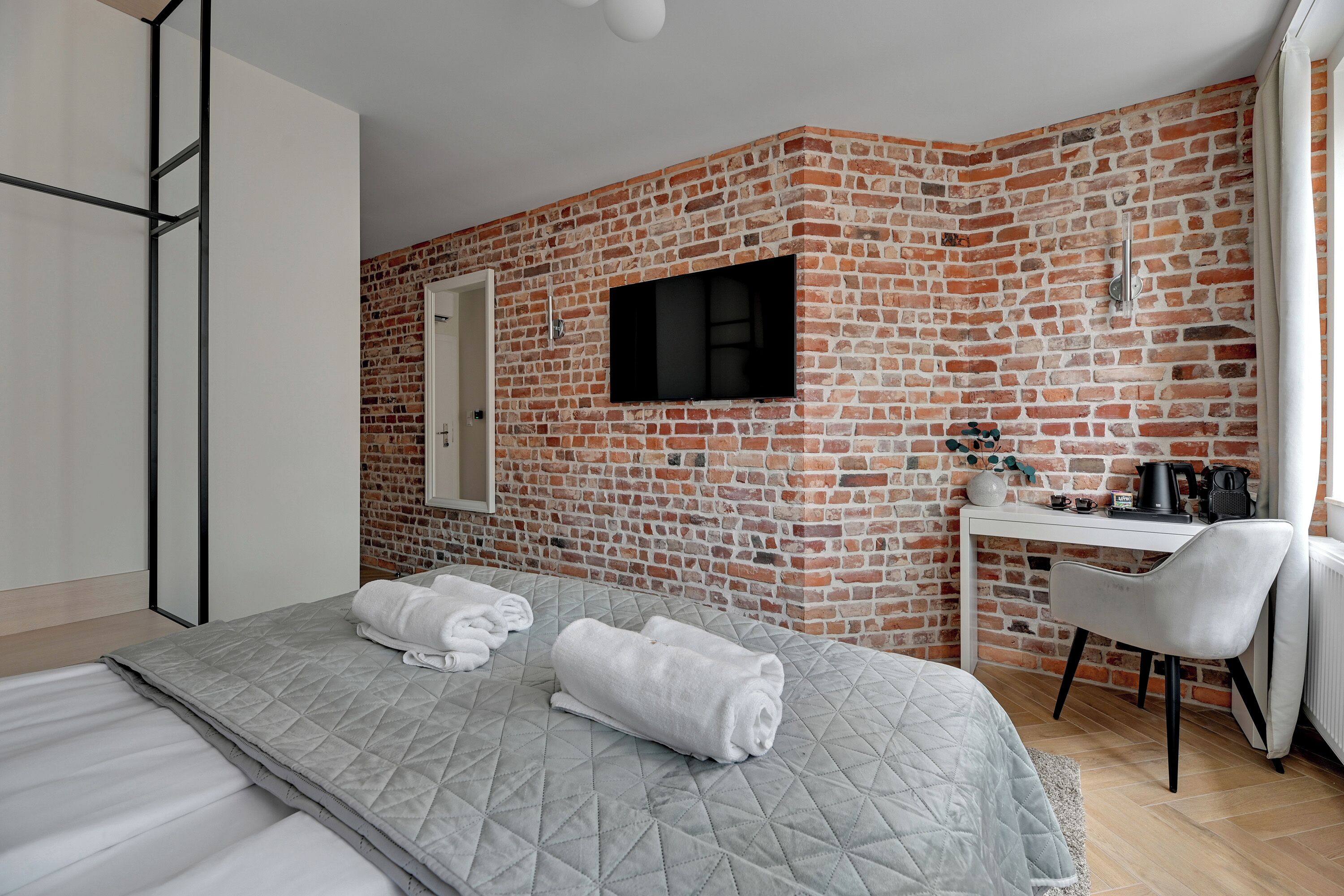 A hotel room with a brick wall, a bed with a quilted grey bedspread, two rolled towels, a white desk with a chair, a flat-screen TV, and a coffee machine.