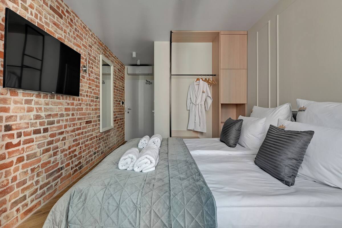 A hotel room with a brick wall, a flat-screen TV, a bed with white bedding, a grey quilted bedspread, and a towel rack with folded towels.