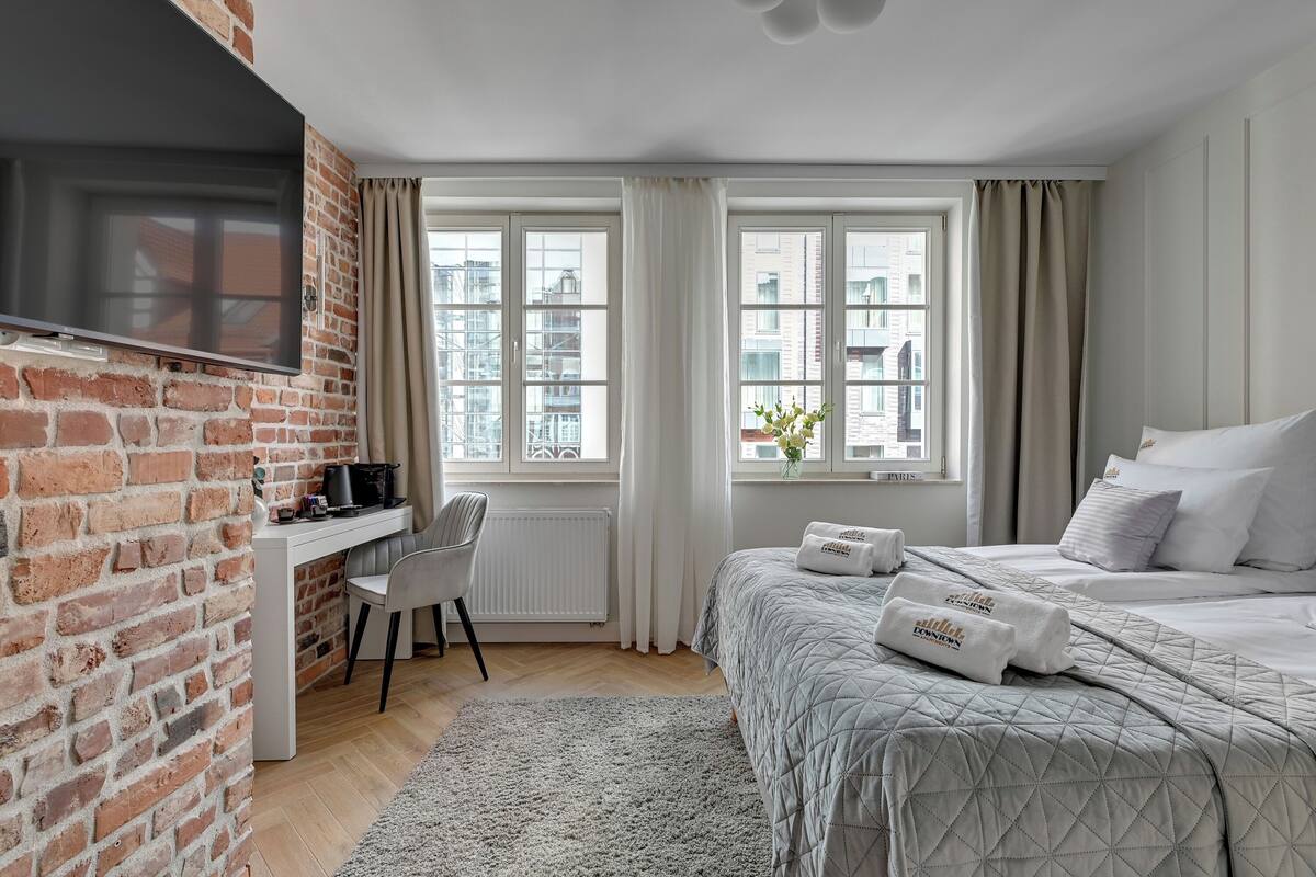 A hotel room with a brick wall, a desk with a chair, a bed with pillows, and large windows with curtains.