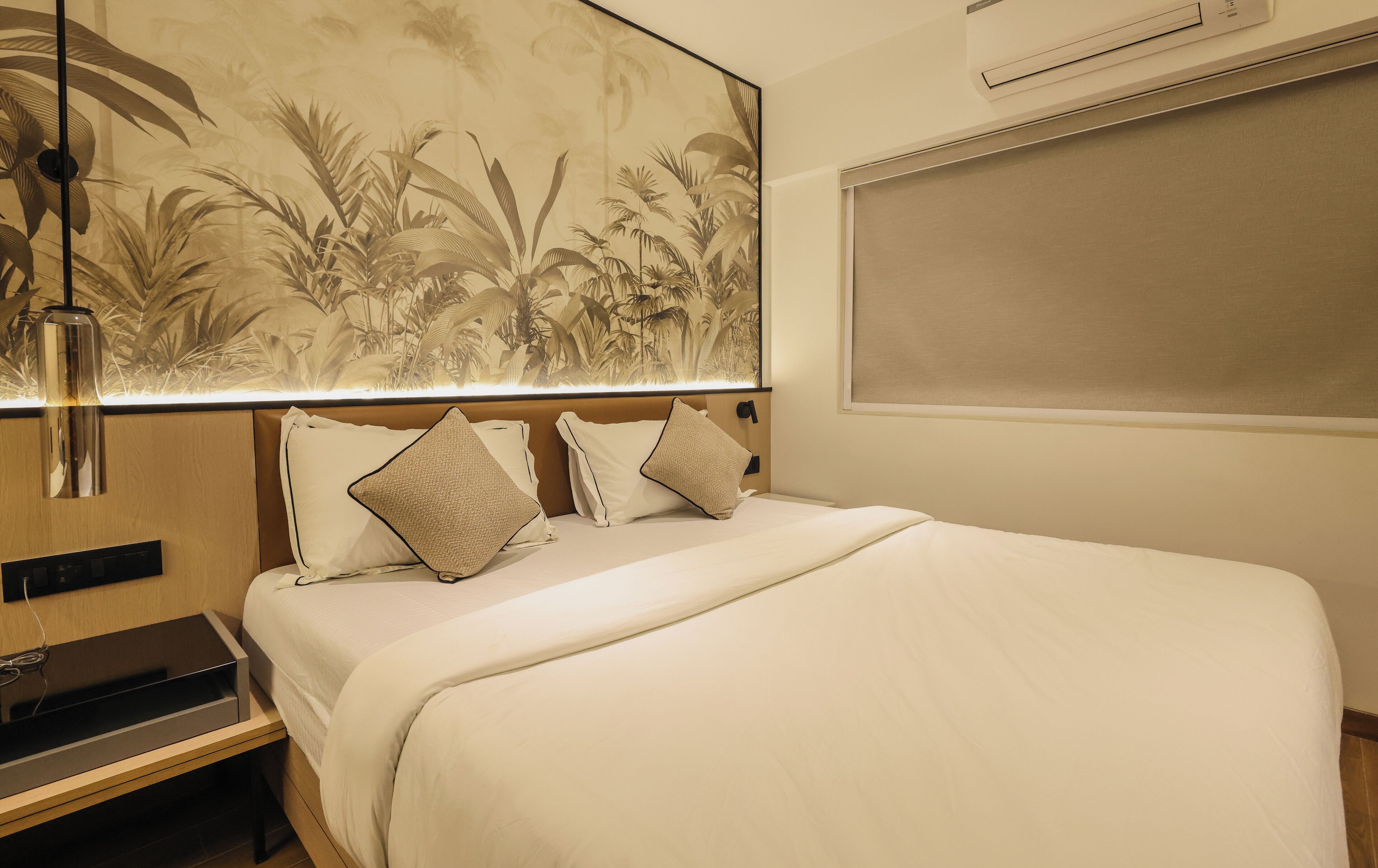 Una habitación de hotel con una cama grande, un cabecero con una lámpara y aire acondicionado instalado en la pared, además de un cuadro de temática tropical sobre la cama.
