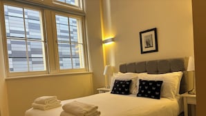 Een net opgemaakt bed met witte lakens en twee decoratieve kussens, een bedlamp en een lijst met een afbeelding aan de muur.