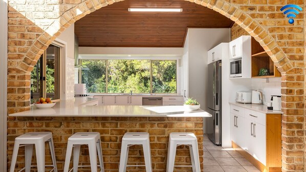 A modern kitchen with a central island, white cabinetry, a stainless steel refrigerator, and a large window offering a view of greenery. - Retreat on Pebble - walk to the beach, a large pet friendly family home (Fingal Bay)