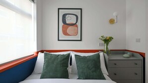 Une chambre d’hôtel avec un lit, des oreillers verts, une table de chevet ornée d’un vase de fleurs et un tableau abstrait encadré accroché au mur.