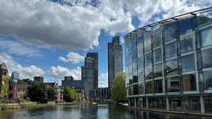 A modern building with large glass windows, a river in the foreground, and a mix of modern and traditional architecture in the background.