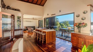 A spacious kitchen with a central island, large windows offering a view of the outdoors, and a dining area with a view of the sea. - Alang Alang, Alang Alang on Hamilton Island (Hamilton Island)