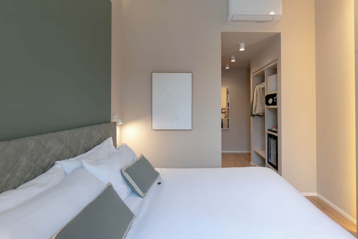Een opgemaakt bed met kussens en een gebogen hoofdbord, een ingelijst beeld aan de muur, en een kast met hangende kleding.