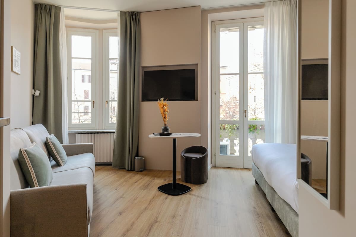 Een hotelkamer met een bank, een bed, een televisie, een klein tafeltje met een vaas, en grote ramen.