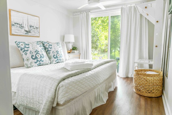A neatly arranged bedroom with a bed, pillows, a bedside table with a lamp, a wicker basket, and a large window with curtains.