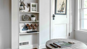 Una habitació amb una porta blanca, una taula rodona amb un llibre i un comandament a distància, i prestatges amb diversos objectes decoratius.