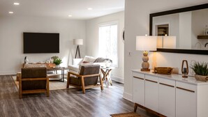 Een moderne woonkamer met een bank, salontafel en een eethoek. Er is een grote flatscreen televisie aan de muur bevestigd, een vloerlamp en een spiegel boven het aanrecht.