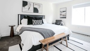 Een moderne slaapkamer met een groot bed, een houten bankje, een kleine ronde tafel en kunstwerken aan de muur.