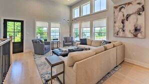 A spacious living room with a large sectional sofa, a patterned armchair, and a coffee table. There are multiple windows providing natural light and a view of greenery outside.