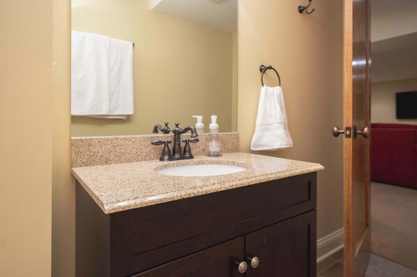 A bathroom with a granite countertop sink, a mirror, a towel rack, and a door leading to a red room.